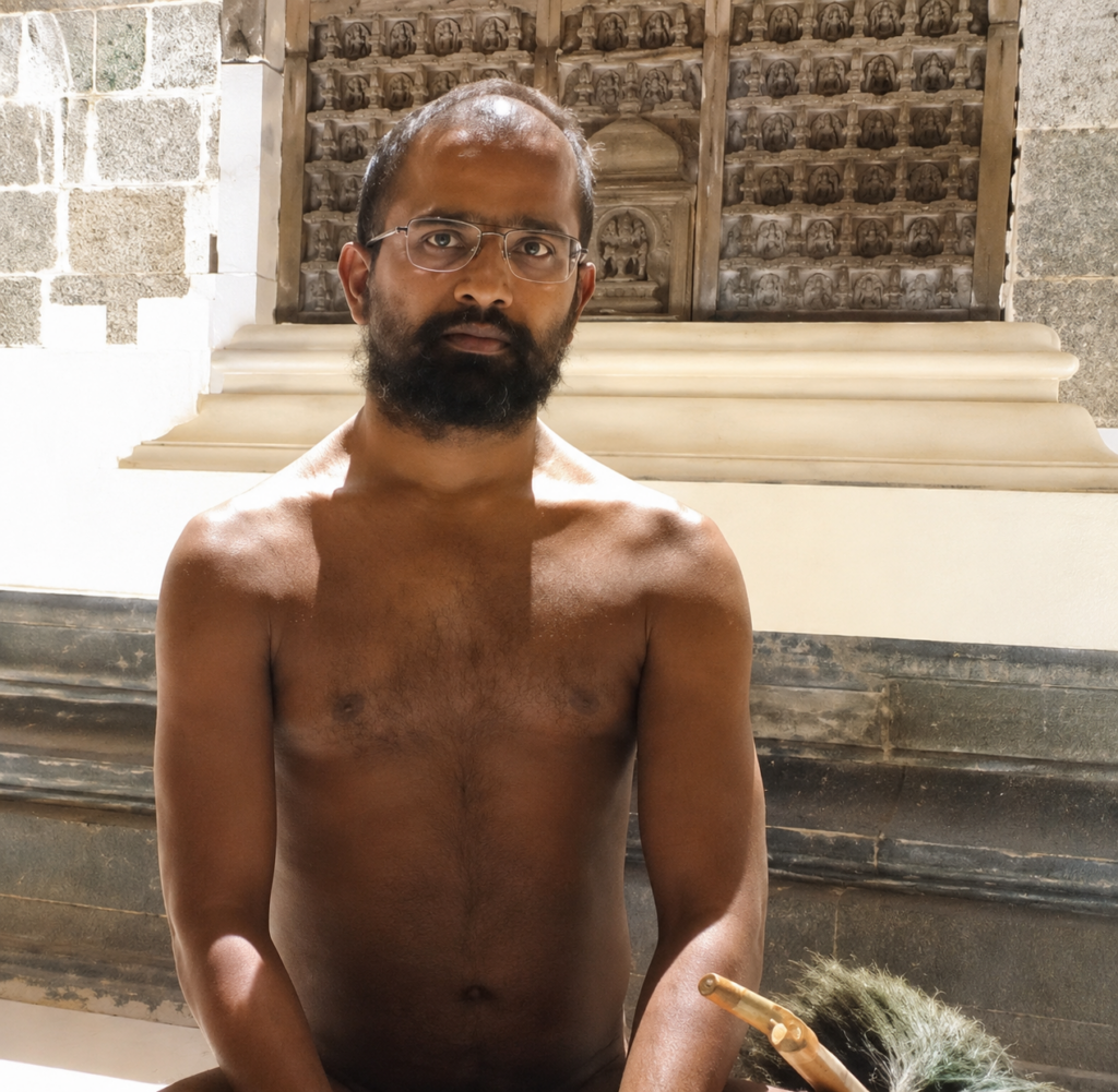 Man with glasses and a beard sits shirtless outdoors in front of a carved stone wall and wooden panel background.
