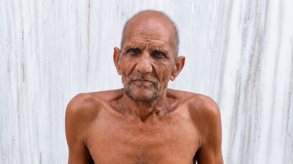 Older shirtless man with a weathered face stands before a white marble wall.