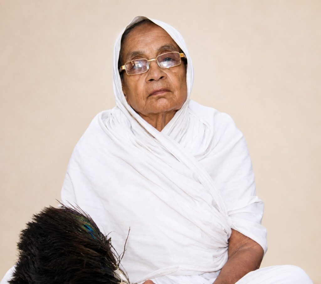 Older woman in a white headscarf and robe, wearing glasses, with a thoughtful expression; peacock feather visible in the lower left corner.