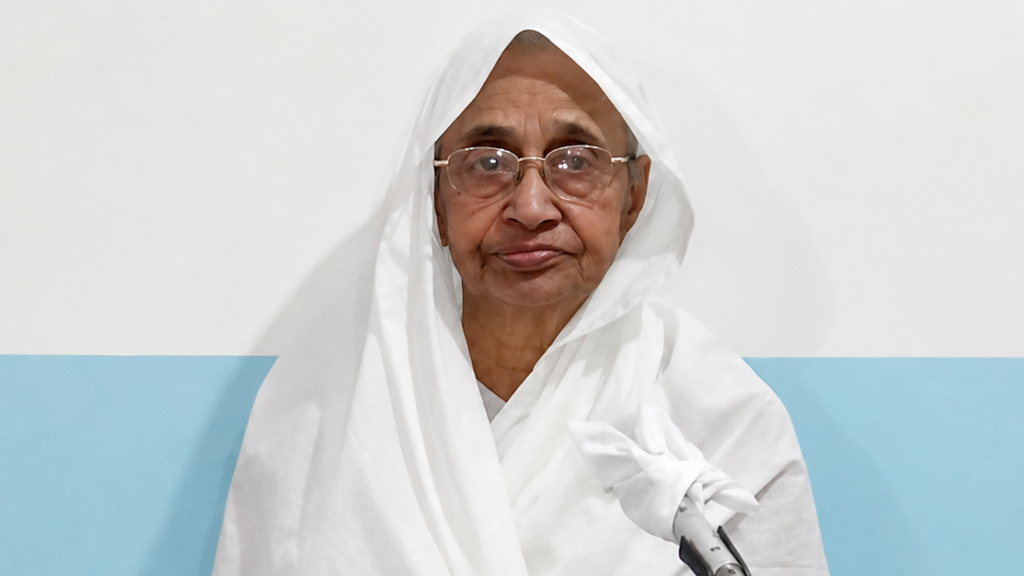 Portrait of an elderly woman wearing a white headscarf and glasses, facing the camera against a white and light blue wall.