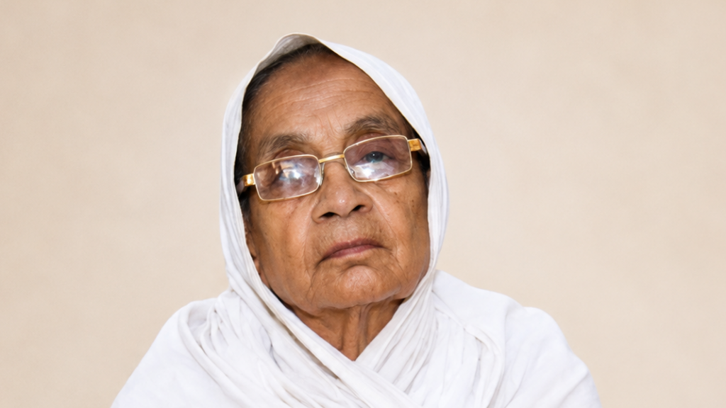 Elderly woman in a white headscarf and glasses gazes pensively at the camera.