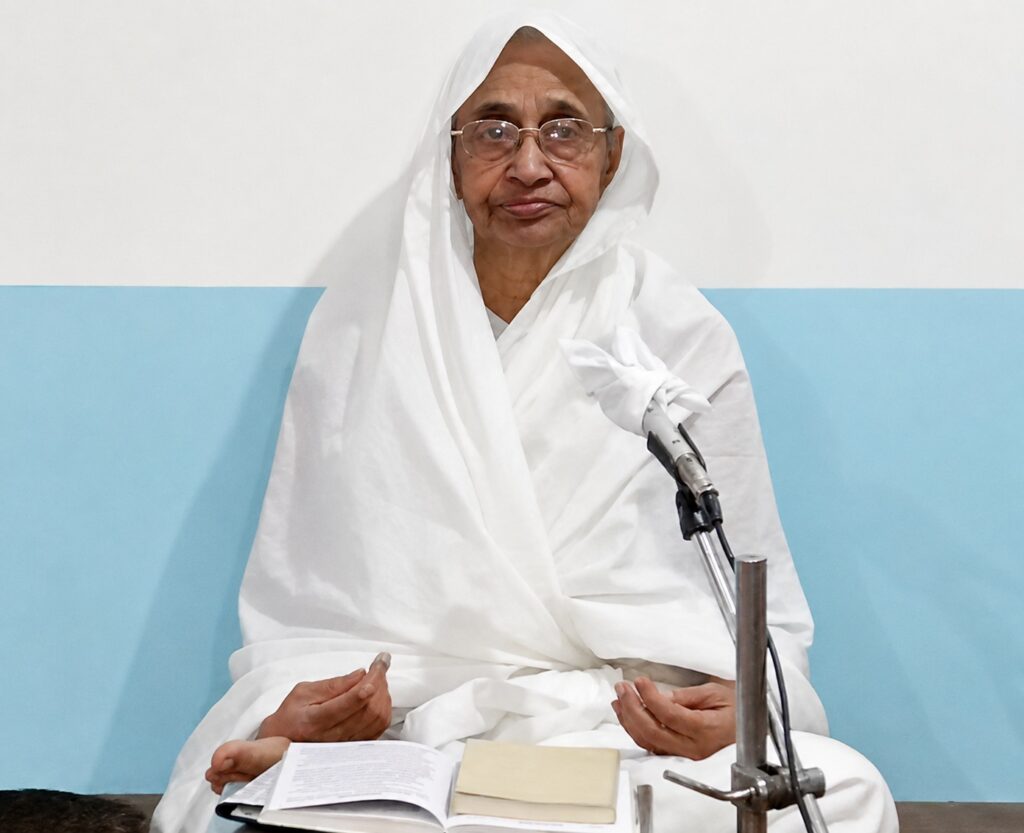 Elderly woman in white robes sits cross-legged, speaking into a microphone with an open book and papers in front.