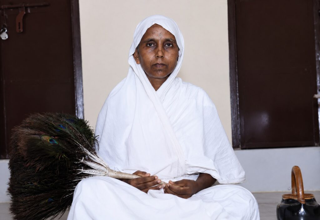 Woman in a white traditional outfit sits cross-legged indoors, holding a peacock feather duster beside her.
