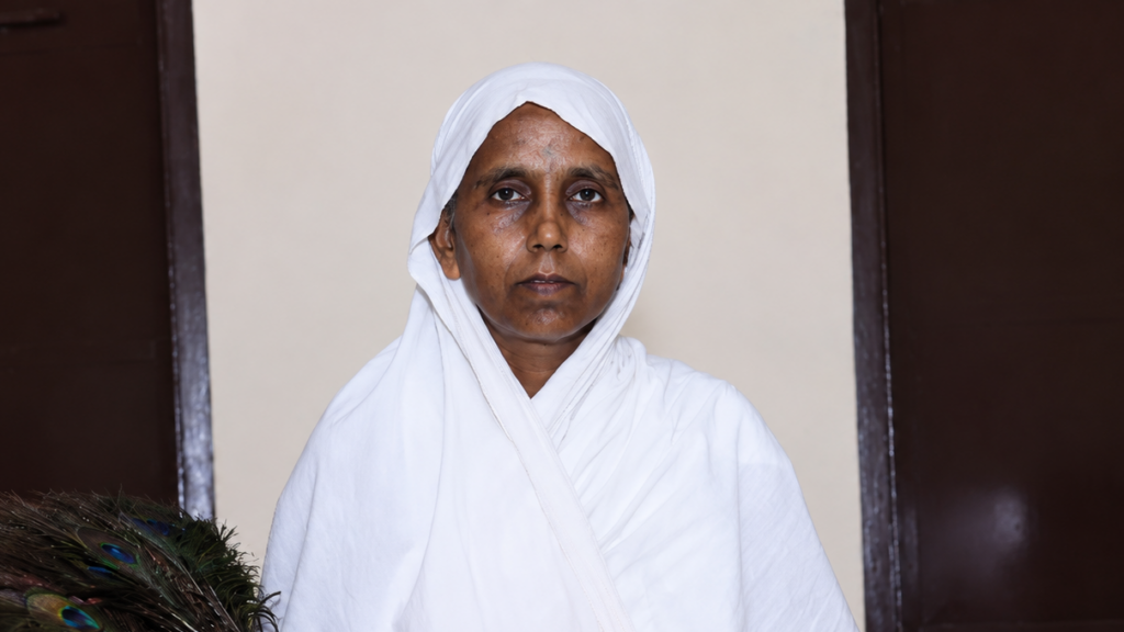 Portrait of a woman in a white headscarf and robe, standing indoors with a calm, direct gaze.