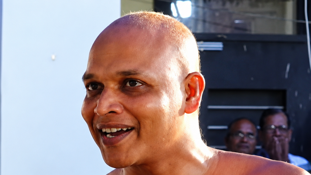 Man with a shaved head smiling outdoors, facing left, with a dark building in the background.