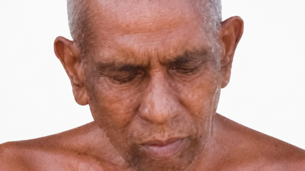 Close-up of an older man with gray hair, shirtless, eyes closed, head bowed in contemplation.
