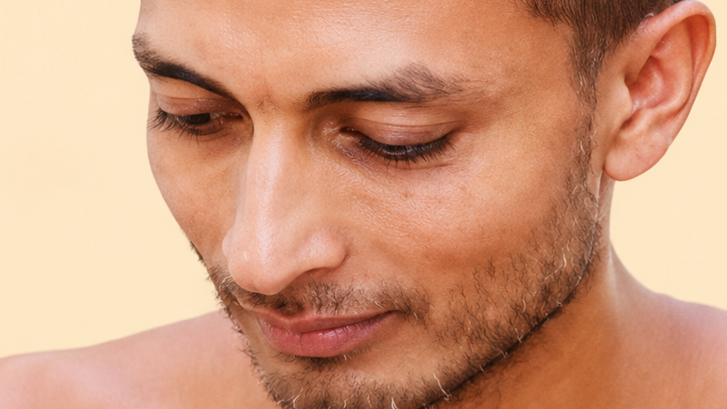 Close-up of a man with short stubble looking downward, showing his jawline and smooth skin on a beige background.