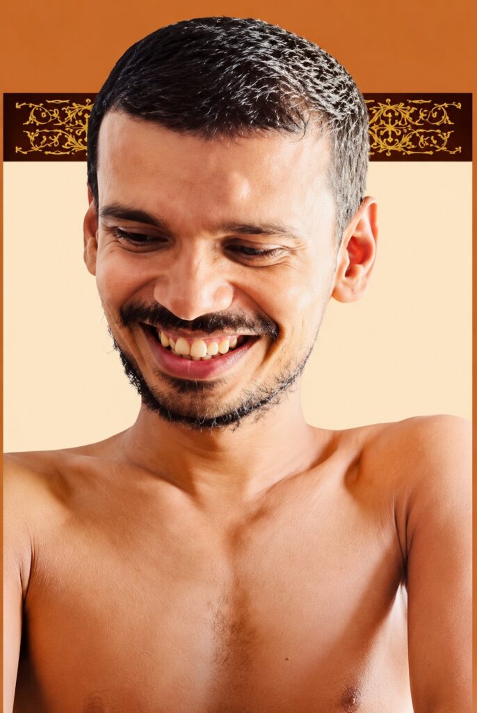 Smiling man with short dark hair and stubble, shirtless against a warm orange background.