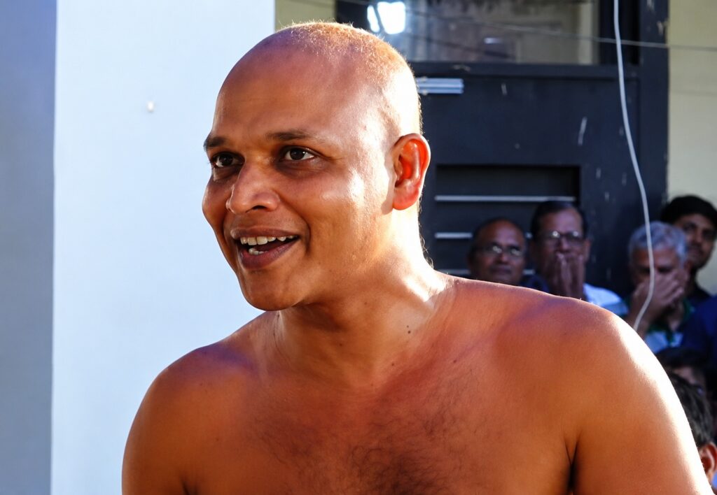 Close-up of a smiling, shirtless man with a shaved head at an outdoor event, audience in background.