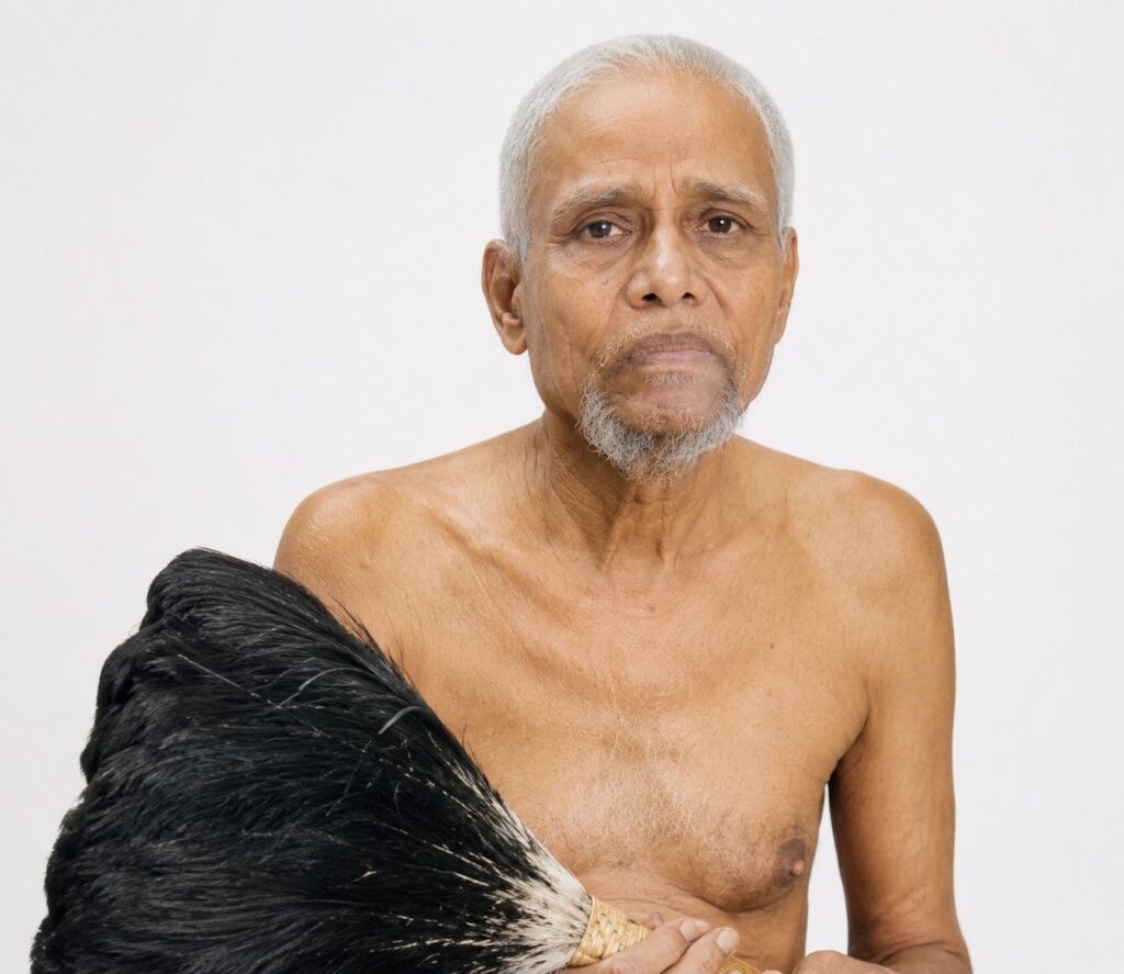 Portrait of an elderly, shirtless man with a gray beard holding a large black-and-white horsehair whisk against a plain white background.