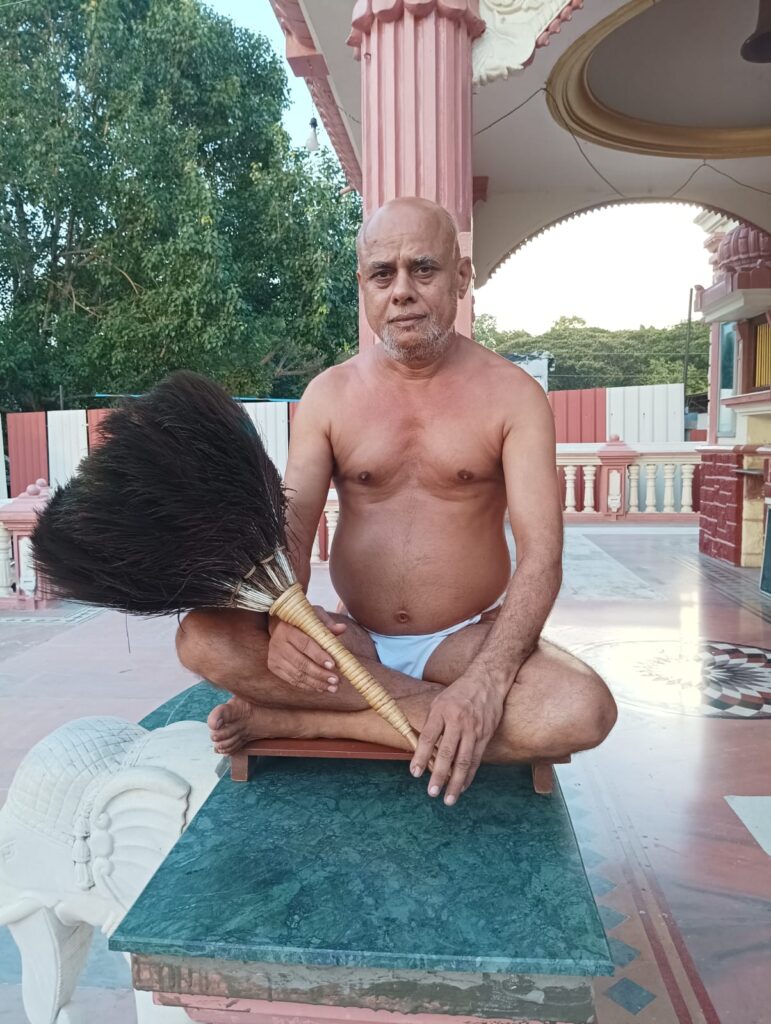 Bare-chested man sitting cross-legged on a small bench, holding a large broom, in a temple courtyard with pink columns.