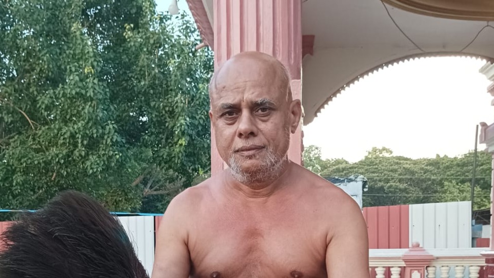 Unidentified man standing shirtless outdoors beside a pink column with green trees in the background.