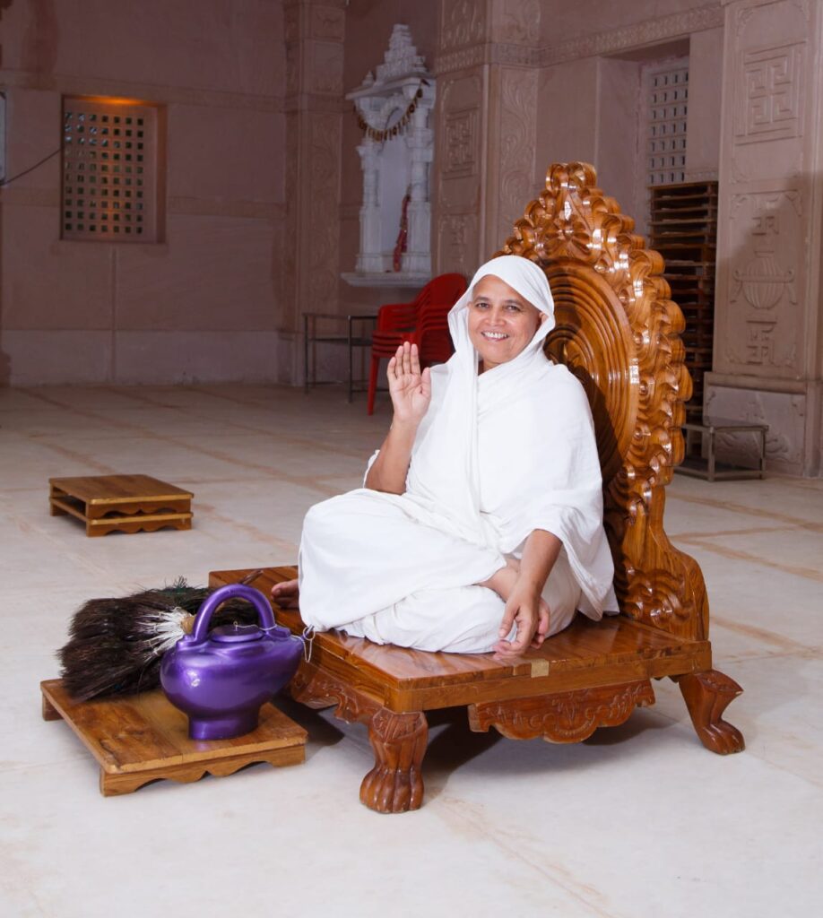 Woman in white robes sits on an ornate carved wooden throne inside a temple, waving smilingly. A purple kettle and broom lie nearby on the floor.