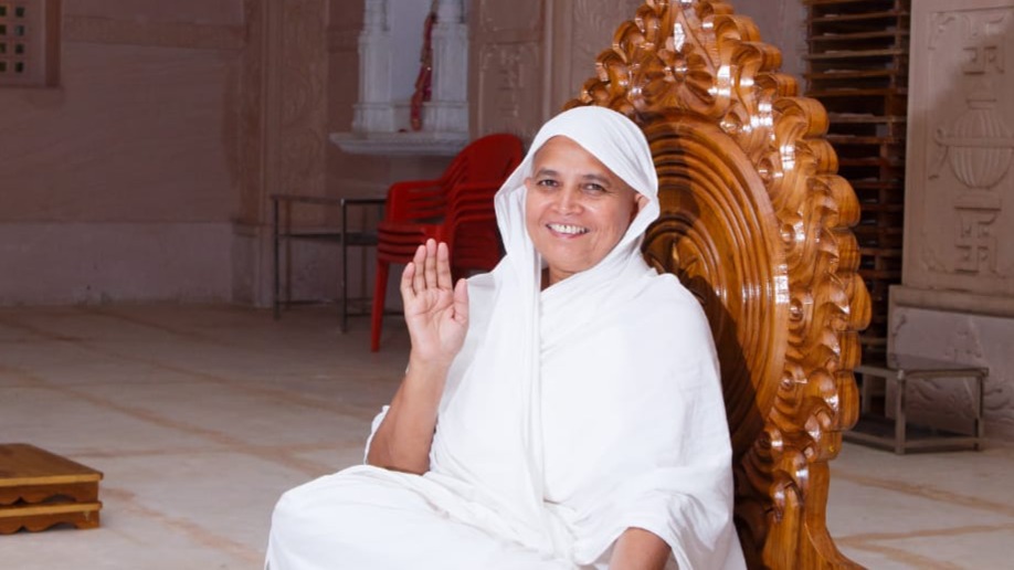 Smiling woman in white robes seated on an ornate carved wooden throne, raising her right hand in blessing.