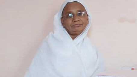 Woman wearing a light blue hijab and glasses, looking toward the camera against a pale wall.