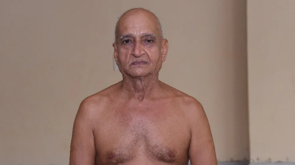 Older, shirtless man standing indoors against a beige wall, looking at the camera with a neutral expression.