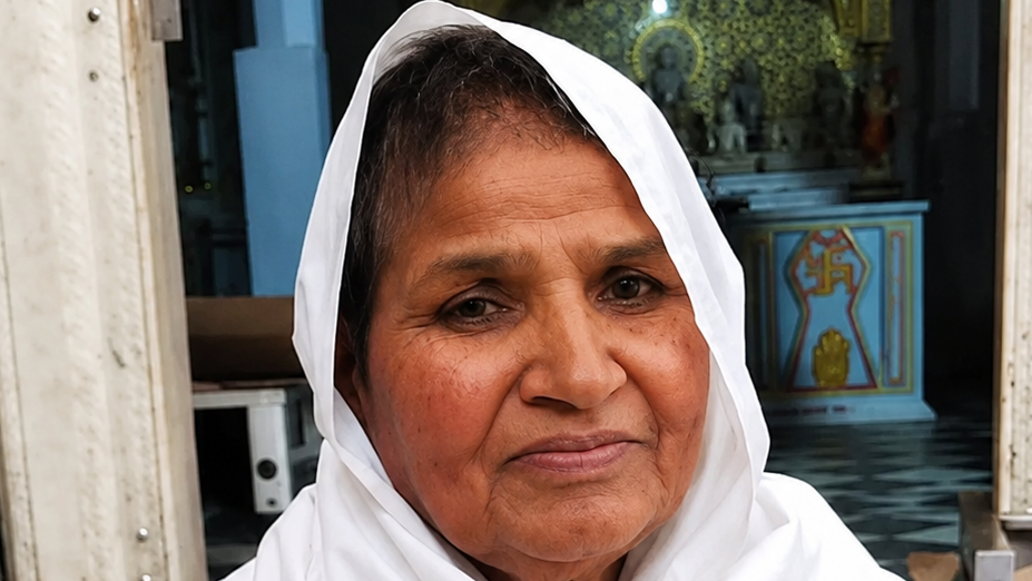 Close-up of a smiling woman with a white headscarf, outdoors near a temple interior.
