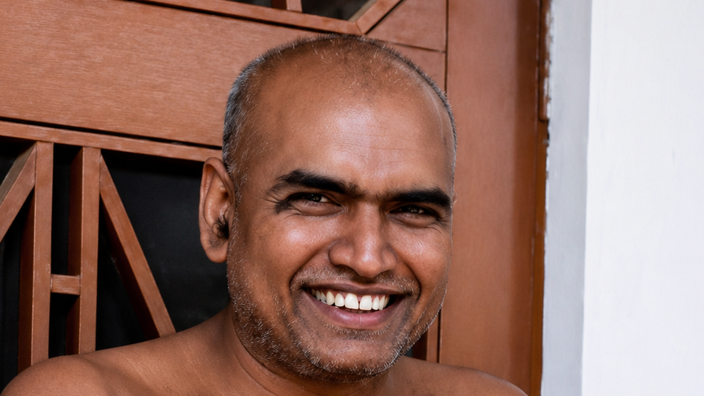 Smiling adult man with short gray stubble, standing near a wooden door frame.