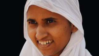 Smiling girl wearing a white hijab against a dark background.