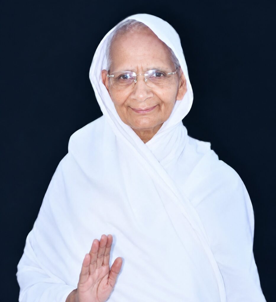 Elderly woman in white headscarf and robe, smiling, raising her right hand in a greeting against a dark backdrop.