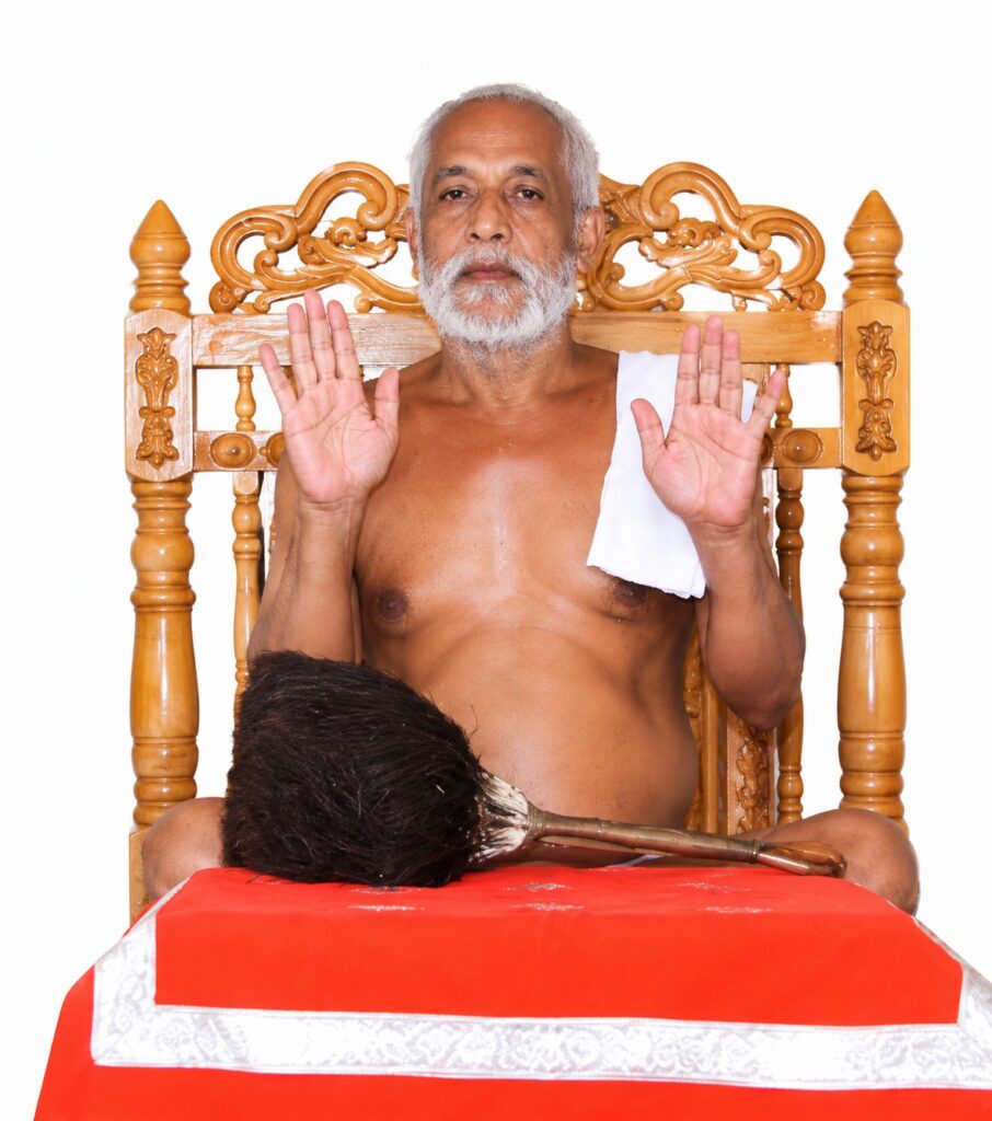 Elder man with gray beard sits shirtless on an ornate wooden throne, hands raised in a calm gesture, white cloth over his shoulder; a person with dark hair rests head on a red cloth-covered surface in front.