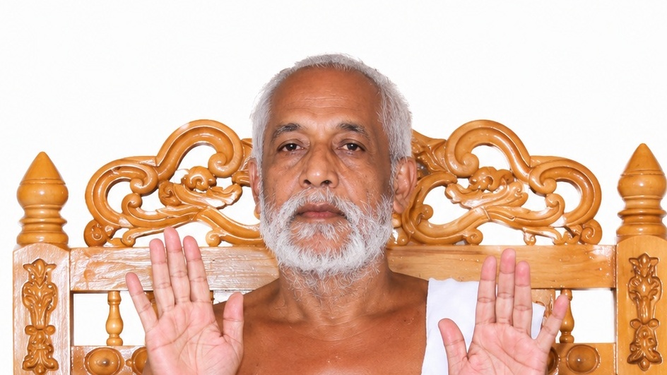 Elderly man with gray hair and beard sits on an ornate carved wooden chair, raising both hands in front of him.