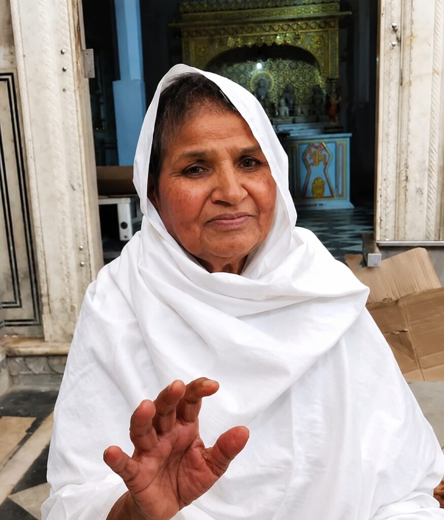 Older woman wearing a white headscarf and robe sits at a temple doorway, hand raised toward the camera.