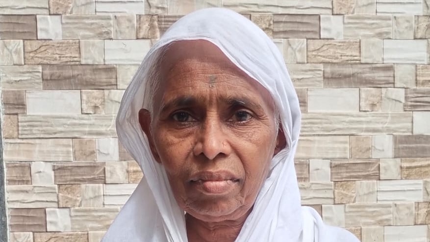 Close-up portrait of an elderly woman wearing a white headscarf, standing in front of a tiled wall.