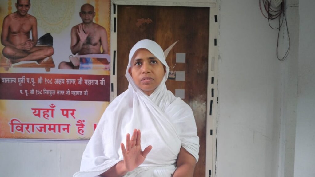 Woman in white traditional attire with headscarf raises her right hand in a stop gesture, standing beside a doorway with a Hindi poster behind her.