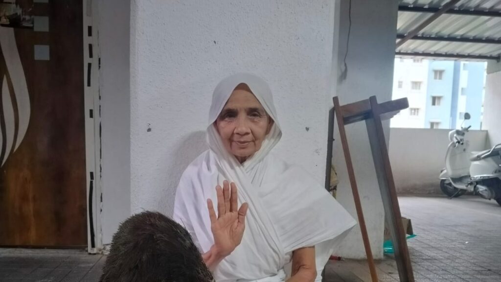 Older woman in a white headscarf sits against a white wall, waving hello to the viewer.