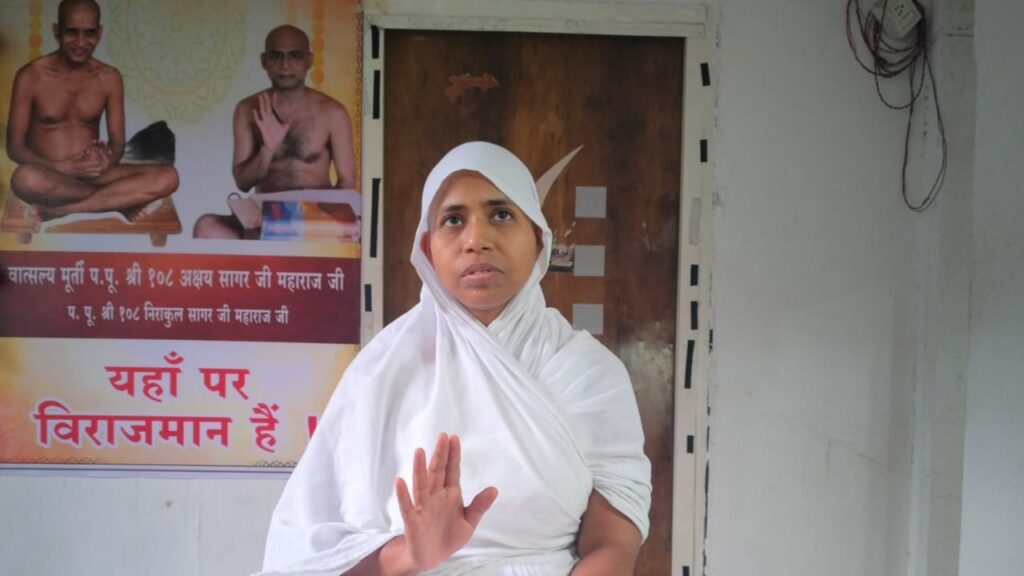 Woman in a white sari and headscarf raises her hand in a stop/hello gesture while seated indoors, with posters on the wall behind her.