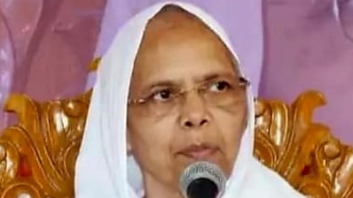 Woman wearing a white headscarf and glasses speaks into a microphone at a podium or stage.