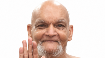 Smiling bald older man with a gray goatee, raising his right hand in a friendly hello.