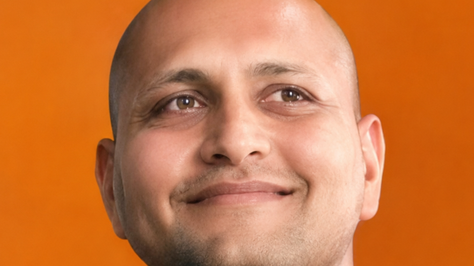 Close-up of a smiling bald man against an orange background with a warm expression and visible stubble on the chin.