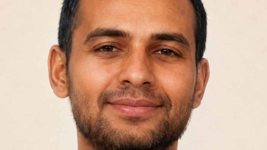 Close-up of a man with short dark hair and light stubble, smiling at the camera against a neutral background.