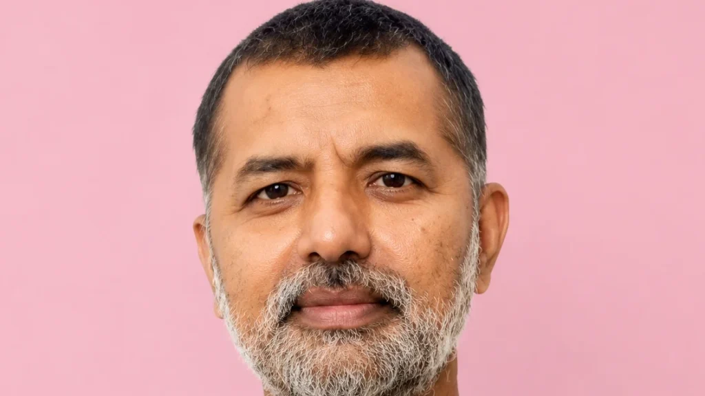 Close-up portrait of a middle-aged man with a gray beard, looking at the camera against a pink background with a calm expression.