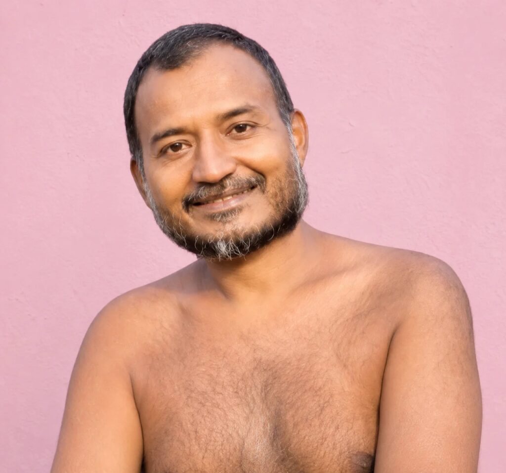 Man smiling at the camera, shirtless, in front of a pink wall.