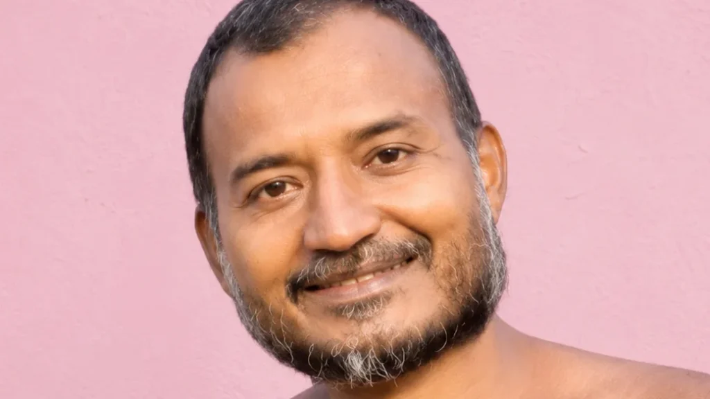 Smiling man with short dark hair and gray beard against a pink wall.