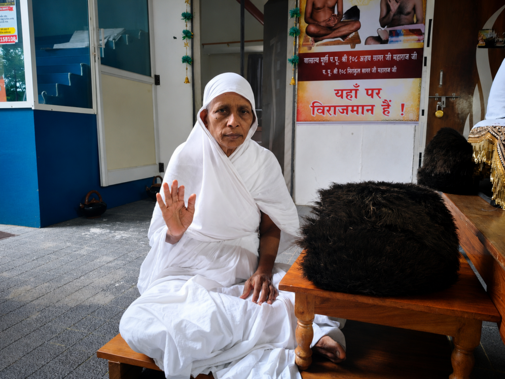 Elderly woman in white robes sits barefoot on a wooden bench outside a building, raising her right hand in blessing; a black fur hat rests on a nearby table.