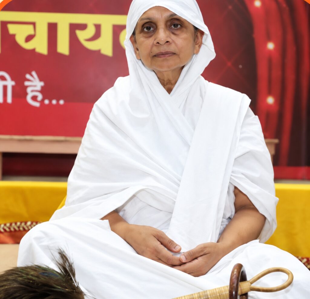 A woman in a white sari sits cross-legged with hands folded in her lap, neutral expression, in front of a red backdrop with yellow text; a wooden ceremonial object is visible in the foreground.