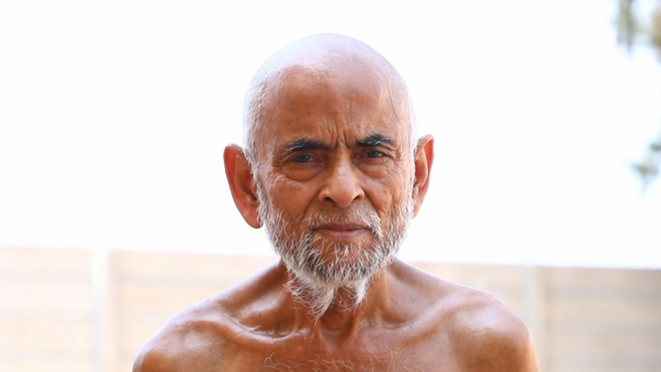 Portrait of an elderly man with a white beard and bald head, bare shoulders, outdoors against a bright sky backdrop.