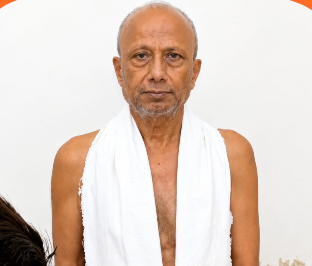 Portrait of an older man with a shaved head and gray stubble, wearing a white towel over his shoulders and looking at the camera.