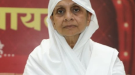 Woman wearing a white headscarf and robe standing in front of a red banner with Hindi text behind her.