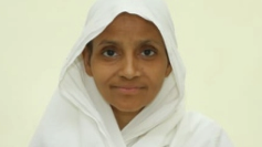 Portrait of a woman wearing a white hijab, smiling at the camera against a pale yellow background.