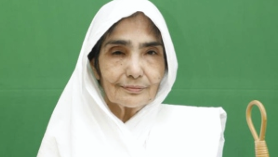 Older woman in a white headscarf and shawl standing with a wooden cane against a green background, facing the camera.