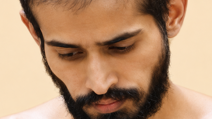 Close-up of a man with a full beard looking downward with a neutral expression on a beige background.