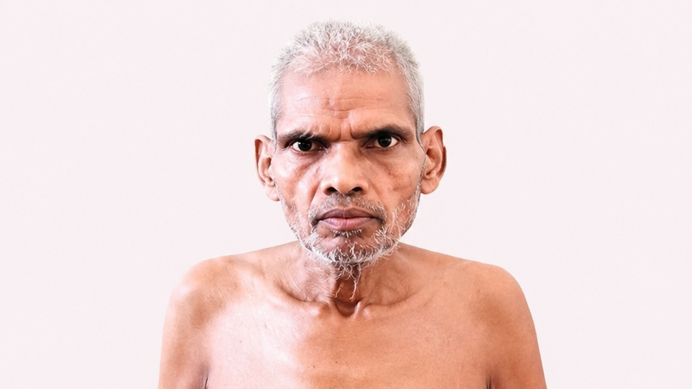 Older shirtless man with gray hair and beard looking directly at the camera against a pale background.