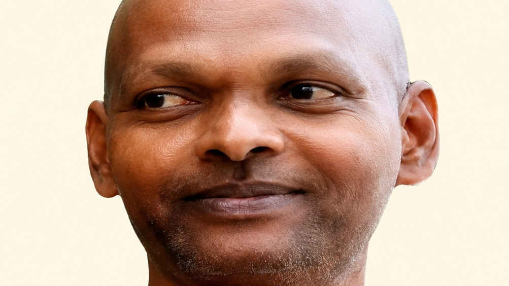 Close-up of a smiling man with a shaved head and light stubble against a pale background, looking slightly to the side.