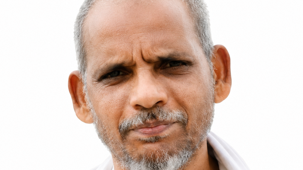 Close-up portrait of a middle-aged man with a gray beard and short hair, squinting slightly with a skeptical expression against a white background.