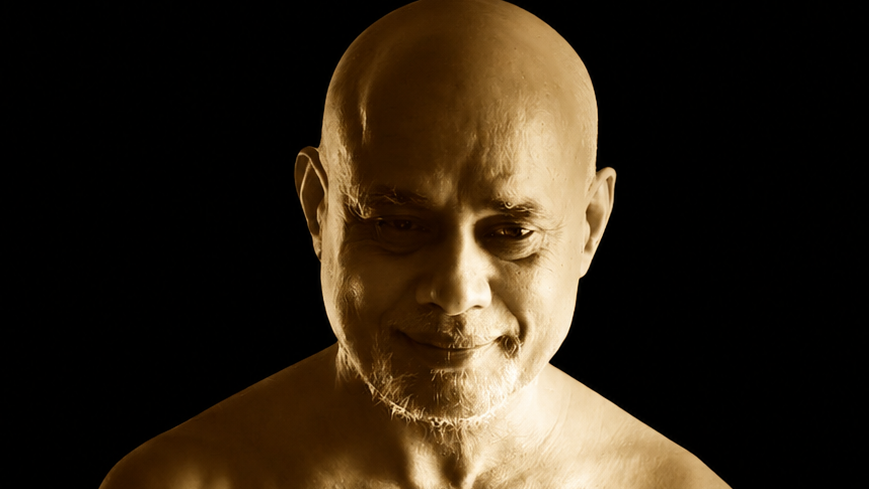 Close-up portrait of a bald man with a light beard, smiling slightly, against a dark background with warm golden lighting.